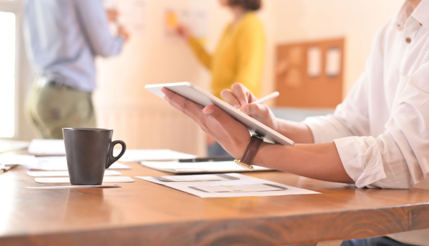 Persona escribiendo en una tablet mientras trabaja en un ambiente colaborativo, con documentos y una taza de café sobre la mesa, y compañeros de equipo debatiendo al fondo. Concepto relacionado con Agencia de SEO en Albacete
