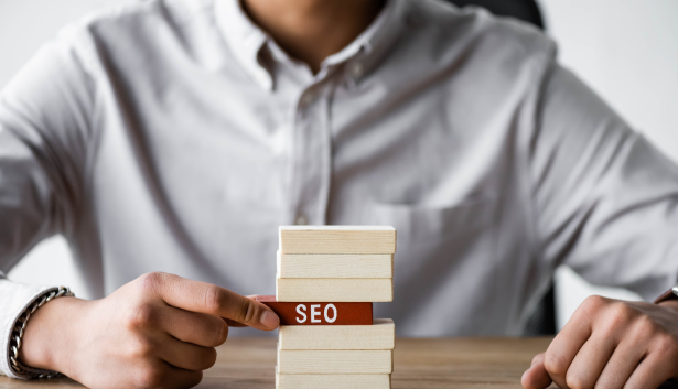 Persona con camisa blanca apilando bloques de madera sobre una pieza roja con la palabra 'SEO' escrita en ella, representando la optimización para motores de búsqueda. Concepto relacionado con Agencia de SEO en Alcorcón.