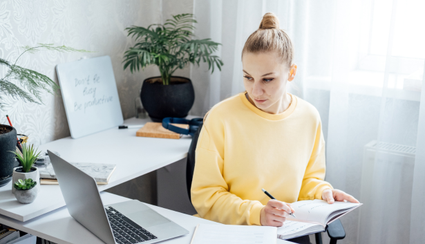 Mujer con sudadera amarilla tomando apuntes en un cuaderno mientras trabaja con una laptop en un escritorio bien iluminado, con plantas y una pizarra motivacional en el fondo. Concepto relacionado con Agencia de SEO en Cartagena.