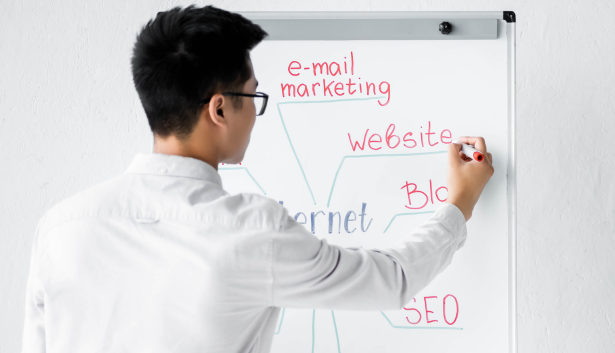 Hombre con camisa blanca y gafas escribiendo en una pizarra conceptos relacionados con marketing digital, incluyendo SEO, blogs, sitios web y e-mail marketing. Concepto relacionado con Agencia de SEO en Elche.