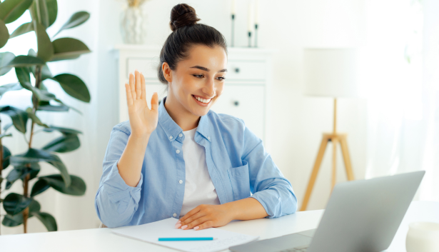 Mujer con camisa azul sonriendo y saludando durante una videollamada en su laptop, representando comunicación digital y trabajo remoto. Concepto relacionado con Agencia de SEO en Fuenlabrada.