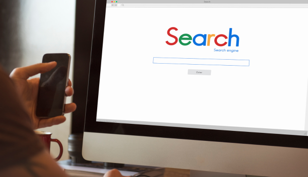 Persona sosteniendo un smartphone mientras observa una pantalla de computadora con un motor de búsqueda abierto, representando estrategias de posicionamiento en buscadores. Concepto relacionado con Agencia de SEO en Huelva