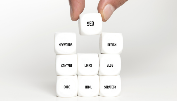 Mano colocando un cubo con la palabra 'SEO' sobre una pirámide de cubos blancos con términos relacionados como 'keywords', 'design', 'content', 'links', 'blog', 'code', 'HTML' y 'strategy', representando los pilares del posicionamiento web. Concepto relacionado con Agencia de SEO en Málaga