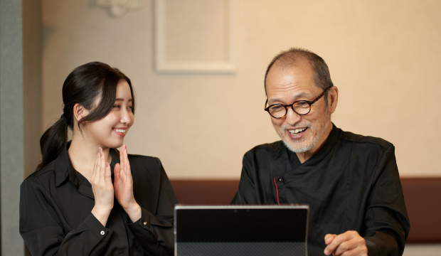 Hombre mayor y joven sonriente vestidos con ropa de chef, viendo una pantalla de computadora con entusiasmo, representando la digitalización de negocios gastronómicos con la ayuda de una agencia de SEO en Vigo.