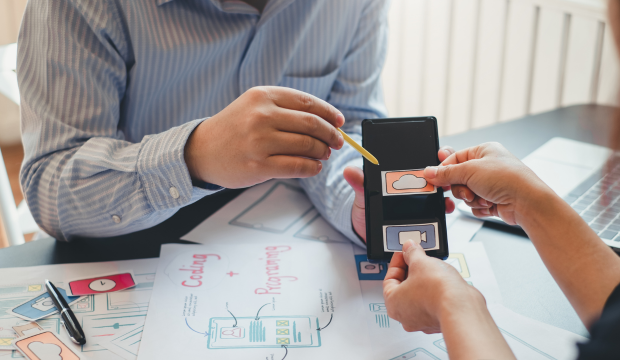 Dos profesionales diseñando una interfaz de usuario para una aplicación móvil, intercambiando iconos de redes sociales sobre un fondo con bocetos y esquemas, reflejando el trabajo estratégico de una agencia de SEO en Vigo.