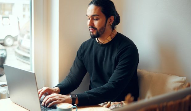 Hombre con barba y cabello largo trabajando en su laptop en una cafetería acogedora, con una taza de café a su lado, representando la flexibilidad y creatividad en el marketing digital de una agencia de SEO en Zaragoza.